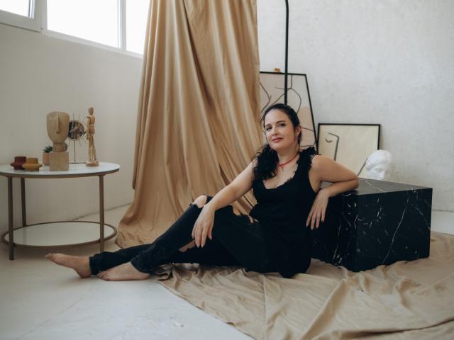 TayaBoret, pieds nus, débardeur noir avec des détails en plumes, pantalon noir, allongée sur un tissu beige, jambes croisées, bras reposant sur une boîte en marbre noir, détendue, souriante, cheveux foncés, collier rouge..