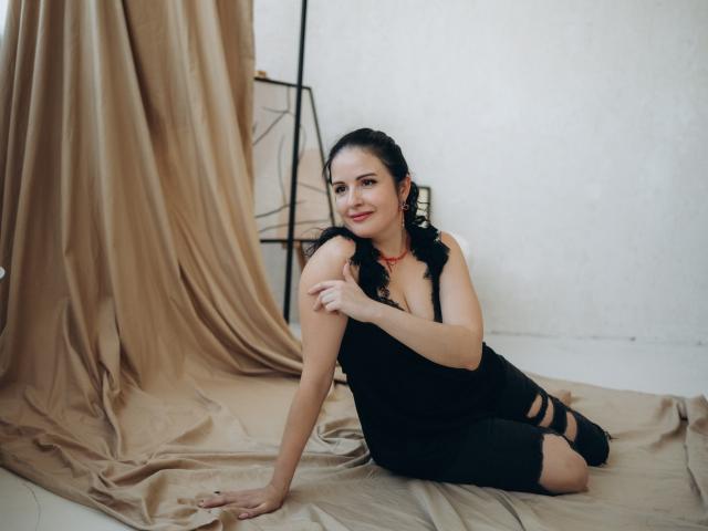 TayaBoret with dark wavy hair, black sleeveless dress, and red necklace, sitting on beige fabric, smiling, hand on shoulder, black strappy shoes, white wall, soft lighting.