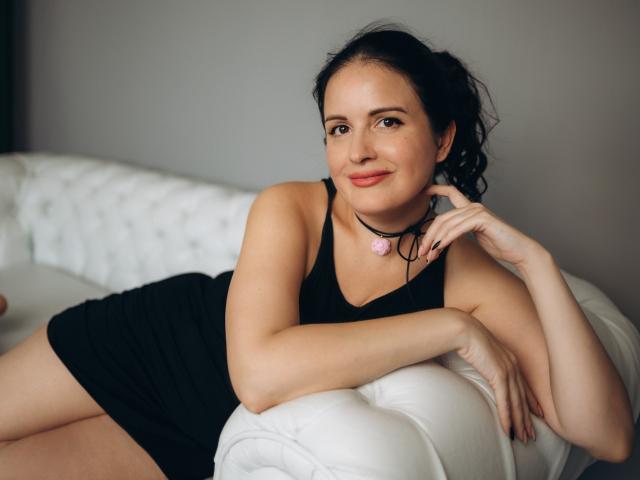 TayaBoret, with curly black hair, lies on a white tufted couch, wearing a black dress, choker with pink flower, smiling, arm resting on couch, hand near chin.