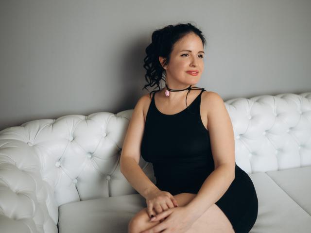 TayaBoret, avec ses cheveux noirs bouclés, sa peau claire et son rouge à lèvres rouge, portant une robe noire moulante et un collier rose, assise sur un canapé blanc capitonné, les mains posées sur ses genoux..
