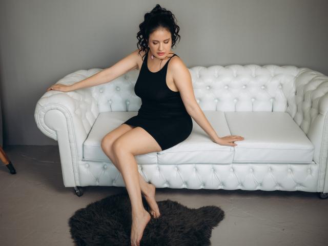 TayaBoret sitting on a white tufted sofa, wearing a black dress, with her ass resting on the seat and her legs crossed, on a brown furry rug.