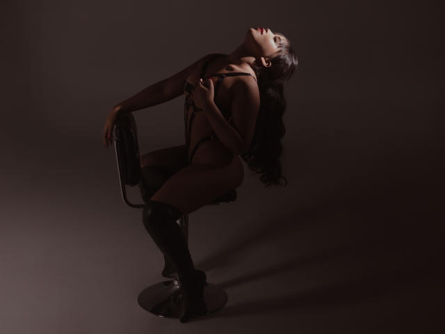 Heyly is bent over with head tilted back, black leather outfit, and stilettos, her hands gripping the chair, moaning.