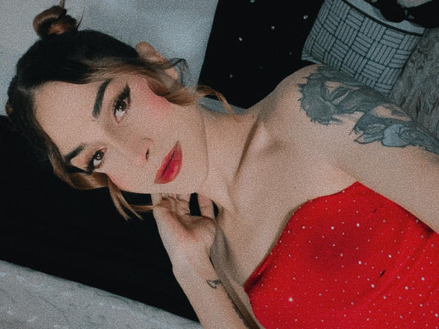 ValentLatin has red sparkly dress, red lips, brown eyes, and a tattoo on her shoulder. Her hair is in a bun, and she's lying on a black pillow.