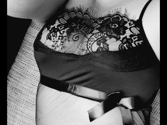 Mistress-lola-hot's chest, showing black lace over dark, smooth fabric with a satin ribbon at the waist, in a black-and-white shot. Boobs are partially covered by lace.