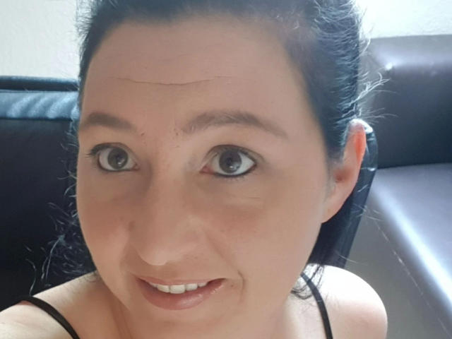 Mistress-lola-hot's face, showing big grey eyes, dark hair in a bun, slight smile, black strappy top, light skin with a small line above her nose.