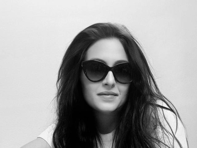 Mistress-lola-hot with long, dark hair, wearing large black sunglasses, slight smile, and white shirt, looking cool and confident in a black-and-white shot.