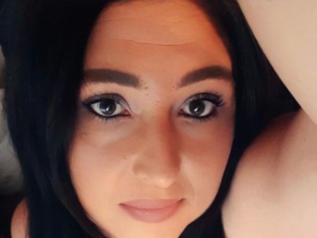 Mistress-lola-hot's face, dark hair, blue eyes, light skin, small smile, subtle makeup, slightly raised eyebrow, visible forehead crease, arm partially in frame, soft lighting, casual selfie.
