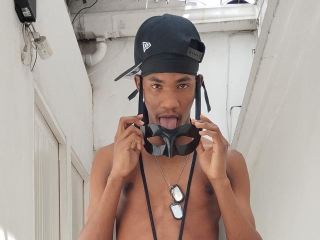 TheCockBoby, shirtless, wearing a black cap, holding a black mask with tongue out, dog tags necklace, standing in a white, narrow hallway, looking at camera.