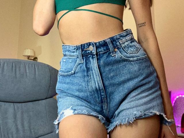 Karish trägt ein grünes Crop-Top, das ihre Brüste zeigt, und eine hoch taillierte Jeansshorts mit fransigen Säumen. Sie hat ein Tattoo am rechten Arm und steht in einem Raum..