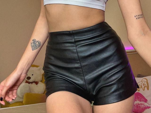 Le short en cuir noir de Karish, qui met en valeur ses fesses musclées et un tatouage à son poignet droit, associé à un débardeur blanc et un tatouage visible sur son bras..