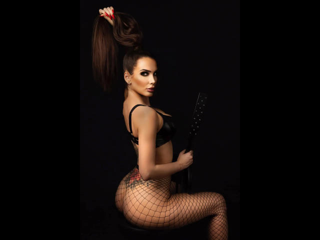 MistressKarolina-hot with huge ponytail, black bra, fishnet pants, holding a guitar, sitting on a stool, sexy, dark background, looking over shoulder, red nails, tattoo on right thigh.