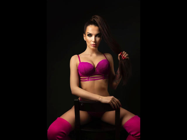 MistressKarolina-hot with pink lace bra and panties, sitting on chair, holding long brown hair, wearing pink thigh-highs, showing her tits and small ass, against dark background.