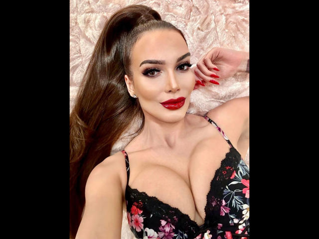 MistressKarolina-hot with huge tits, wearing a floral black lace bra, red lipstick, long brown hair in a ponytail, and red nails, lying on a pink fluffy surface.