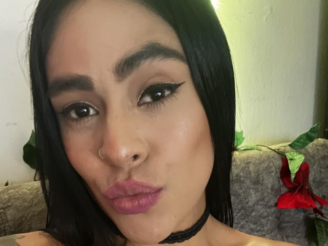 StellaBeckker with black hair, brown eyes, winged eyeliner, pink lipstick, nose ring, black choker, pouty lips, and a slightly confused expression. Her tits are not visible.