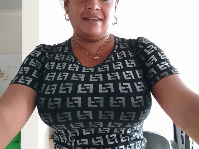 Crysmia with medium brown skin, smiling, wearing a black geometric patterned top, gold necklace, and earrings, showing her large tits and curvy ass in a selfie.