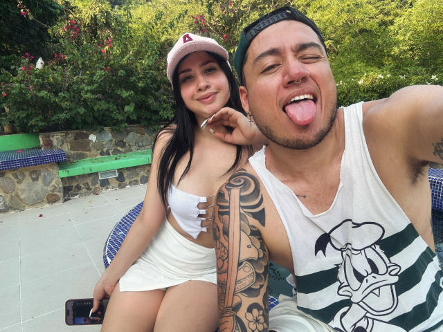 LucyAndJake with black hair, small tits, white crop top, and shorts, sitting next to Jake, who's sticking out his tongue, tattooed arm, white tank, and green striped shirt.
