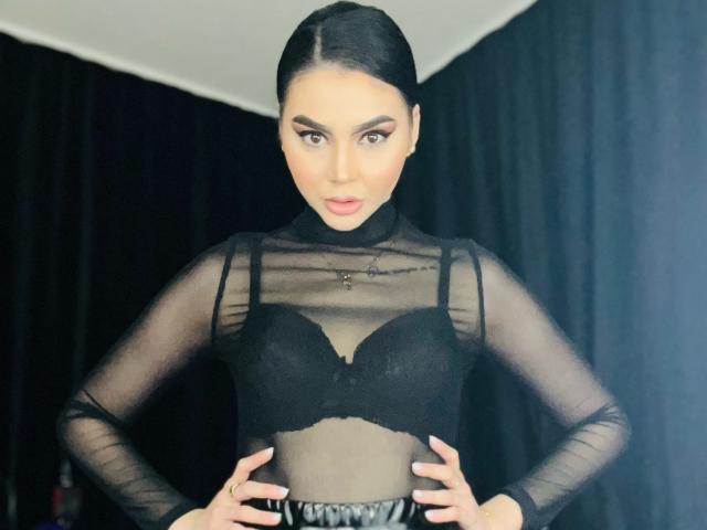 MistressEdithFuchs with black bra, sheer long-sleeve top, hands on hips, dark hair slicked back, serious expression, black curtains in background, focused gaze, subtle makeup.
