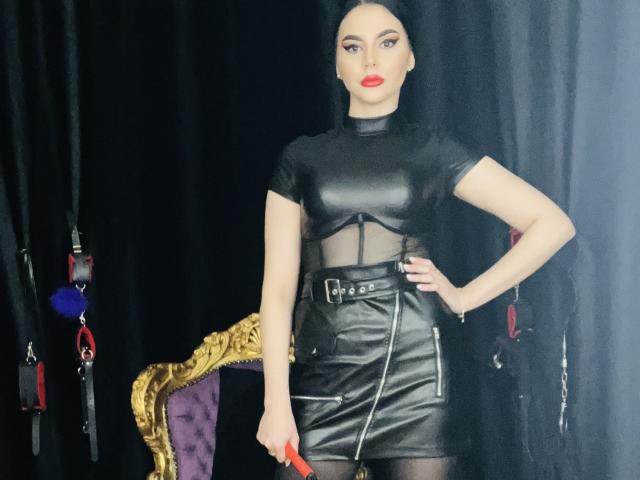 MistressEdithFuchs stands with a hand on her hip, wearing a black leather dress with mesh, holding a red paddle. She has red lips, black hair, and a confident look.