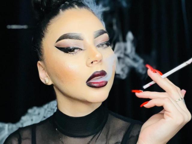 MistressEdithFuchs met smoky oogmake-up, opvallende rode lipstick en donkerrode nagels, die een sigaret vasthoudt, een doorschijnend zwart top draagt en een zelfverzekerde, sexy uitdrukking heeft..