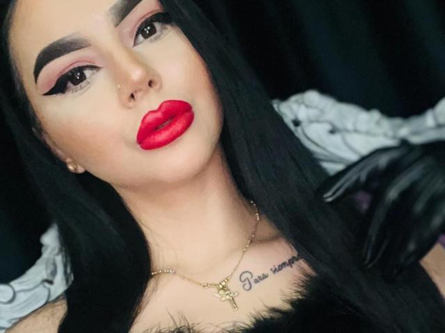 MistressEdithFuchs with red lips, black eyebrows, and long black hair, wearing a gold necklace with a cross, has a tattoo above her tits. She's looking at the camera.