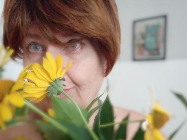 MioritaStar with short red hair, blue eyes, and fair skin, holding yellow flowers partially covering her face, smiling slightly. Her tits are not visible, focus is on face.
