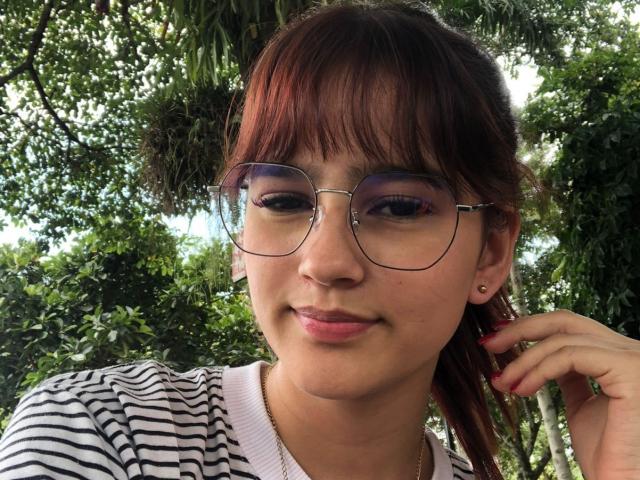 MelodyMansson has red-brown hair, glasses, and a cute smile. She's wearing a striped shirt with a gold necklace, and her hand touches her ear. She's looking at the camera.