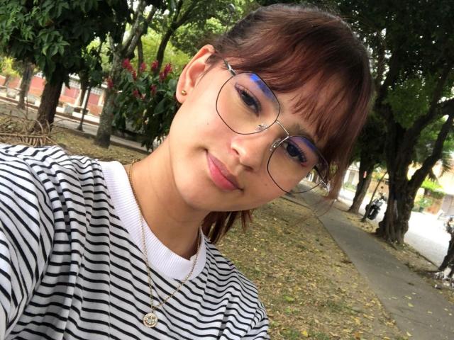 MelodyMansson has brown hair, blue eyeshadow, and wears round glasses. She's smiling, wearing a striped shirt, and a gold necklace. Her tits are small, and she's cute.