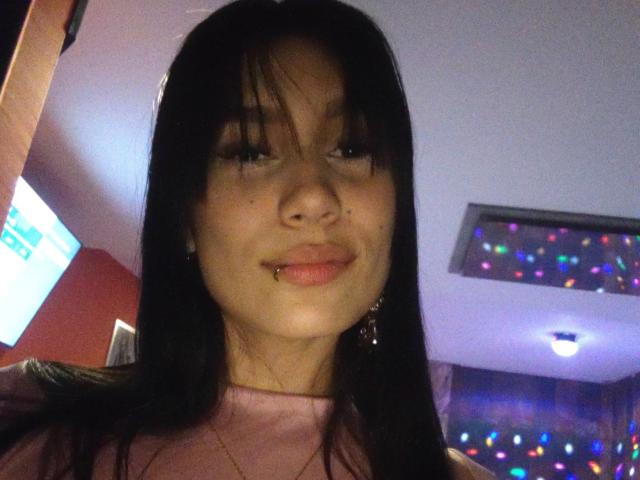KataleyaMansson with black hair, slightly parted lips showing a labret piercing, wearing a pink top, and a gold necklace, in a dimly lit room with colorful lights.