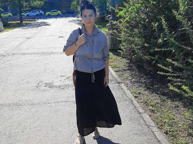 LolaGreate stands on a sunlit sidewalk, wearing a gray button-up shirt, black skirt, and sandals. She's got small tits, dark hair in a bun, and a shoulder bag.