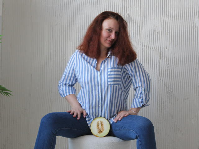 BarbaraStily, avec ses cheveux roux bouclés, portant une chemise bleue à rayures et un jean bleu, est assise les jambes écartées, une tranche de pastèque posée sur son entrejambe, les mains sur ses cuisses..