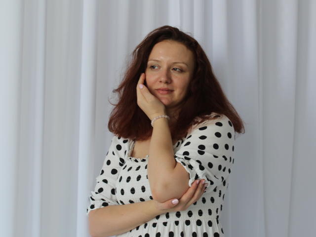 BarbaraStily with wavy brown hair, white polka-dot dress, touching cheek with hand, subtle smile, pale skin, light makeup, white curtain background, bracelet on wrist.