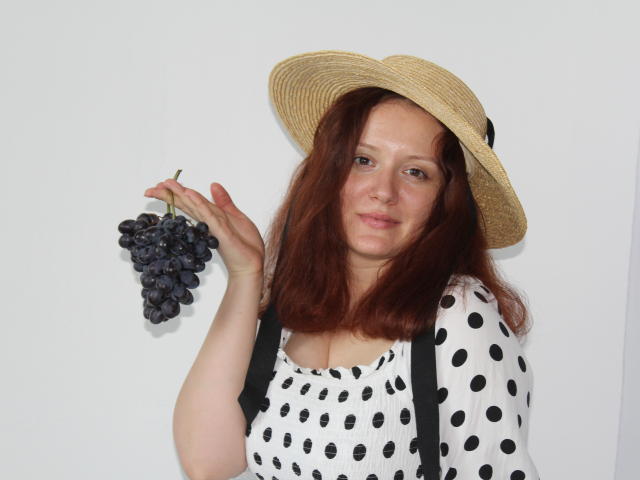 BarbaraStily met rood haar, die een strohoed draagt, een tros paarse druiven vasthoudt, een jurk met stippen, zwarte bretels, lachend, witte achtergrond..