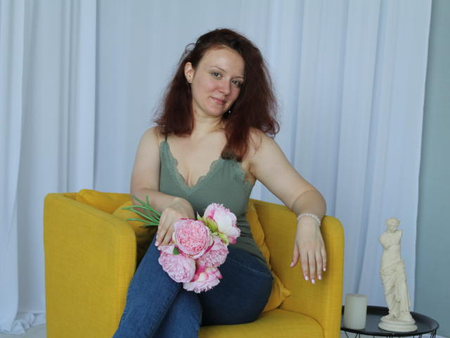 Barbara, avec des cheveux bruns ondulés, un débardeur en dentelle vert, un jean bleu, tenant des pivoines roses, assise sur une chaise jaune, avec un rideau blanc, une petite statue et une bougie blanche en arrière-plan..
