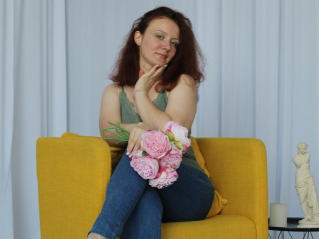 BarbaraStily with pink peonies, sitting on yellow couch, green tank top, blue jeans, hand on chin, smiling, brown wavy hair, white curtain background, small statue on right.
