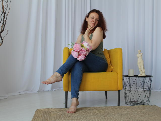 BarbaraStily, avec ses cheveux bruns bouclés, un débardeur vert, un jean bleu, pieds nus, tenant des pivoines roses, assise sur une chaise jaune, une petite table noire avec une statue blanche et une bougie..