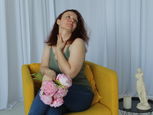 BarbaraStily with reddish-brown hair, green tank top, and blue jeans, holding pink peonies, sitting on yellow chair, hand on neck, white curtain backdrop, small statue on table.