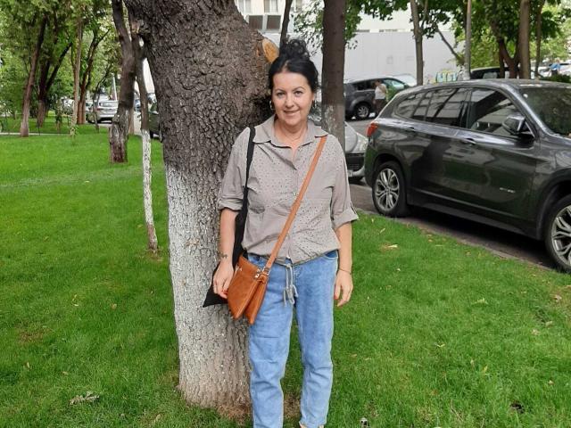 LolaGreate debout contre un arbre, portant une chemise grise, un jean bleu, un sac bandoulière marron, et les cheveux noirs attachés en chignon, avec un fond d'herbe verte et une voiture grise..
