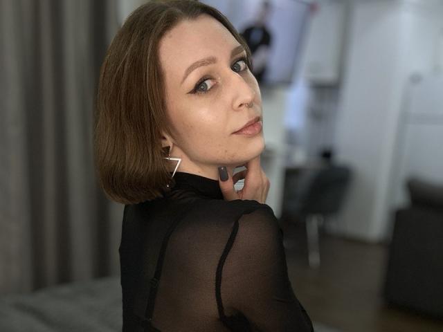 OaklynDobson with short brown hair, black sheer top, and triangle earrings, looking back with a slight smile, nose ring, and black eyeliner. Oaklyn's ass is partially visible.
