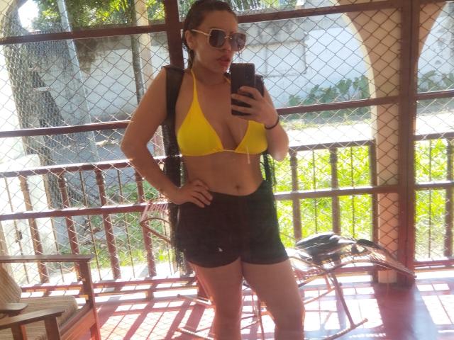 VioletaStone with tan skin, brown hair in a bun, wearing a yellow bikini top, black shorts, sunglasses, and a backpack, standing in a sunlit, fenced, wooden, outdoor area.