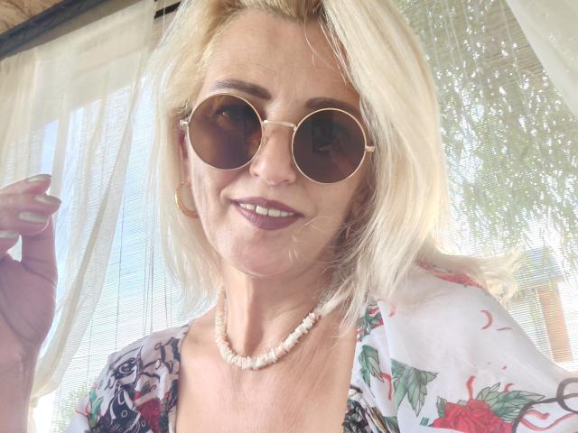 SexyCoco, avec des cheveux blonds, portant des lunettes de soleil rondes, un haut fleuri, un collier de perles et du rouge à lèvres foncé, souriante, tenant un rideau, montrant légèrement ses seins, en plein jour..