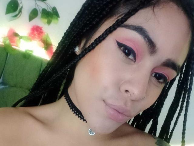 StellaBeckker, com cabelo trançado, sombra rosa, delineador preto, piercing no nariz, colar preto com uma lua crescente e um sorriso discreto, mostrando seus ombros nus..