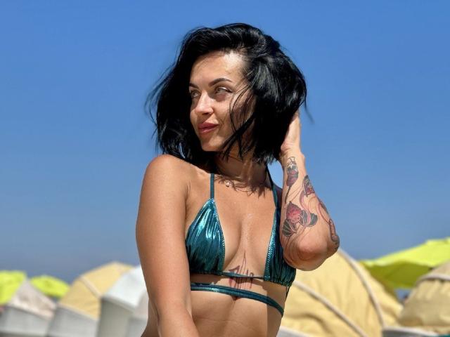HollyHope with short black hair, green metallic bikini top, tattoos on right arm, standing under blue sky, hand in hair, smiling, showing small breasts, sunny, beach.