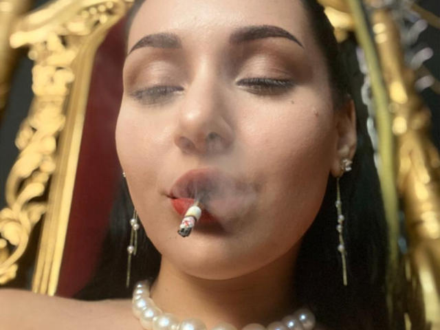 LexyClaire with her eyes closed, a cigarette in her mouth, pearl necklace, and earrings, showing off her slightly flushed, smoky face and dark hair.