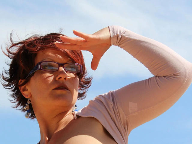 Popp-Sylvie-hot with short red hair, glasses, and a light pink shirt, raising her right arm. Her tits are partially visible, and she's wearing colorful earrings.