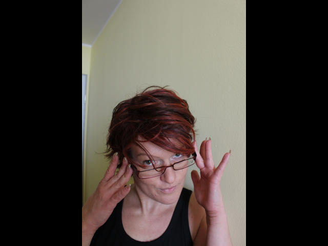 Popp-Sylvie-hot with short red hair, wearing glasses, black tank top, hands near face, smiling slightly. Popping glasses off with fingers. Photo.