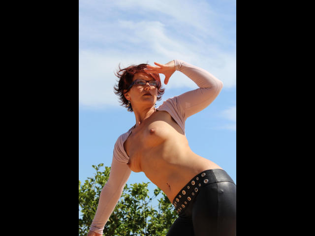 Popp-Sylvie-hot with short brown hair, glasses, and a tan skin tone, wearing a pink cropped top and black leather pants, exposing her small tits and flat ass, against a blue sky.