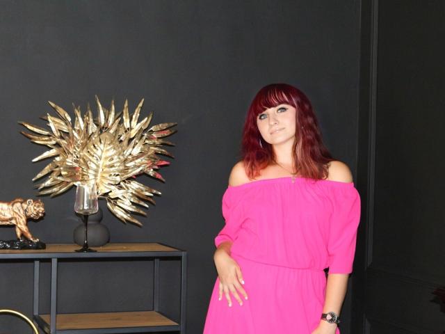 NinaFlawlees with red hair, off-shoulder pink dress, standing, black wall, gold spiky lamp, bronze tiger statue, and black shelf in the background. Her tits are partially visible.