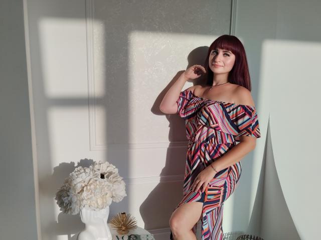 NinaFlawlees with red hair, wearing an off-shoulder, colorful, patterned dress, standing in sunlight, touching her chin, near a white vase with white flowers and a gold sunburst.