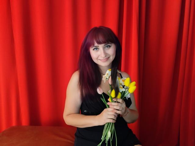 NinaFlawlees with red hair, holding yellow and white flowers, smiling, wearing a black dress, sitting on an orange chair, in a red curtain setting.