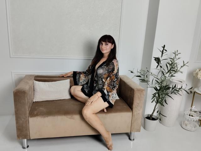 NinaFlawlees sits on a beige sofa, wearing a floral kimono, exposing her bare legs and pussy, with dark hair, and a small pillow beside her.